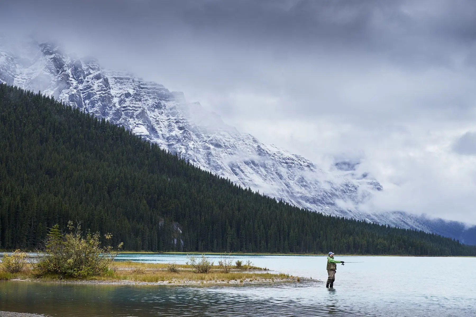 An angler casting from the shallows of a turquoise mountain lake beneath snow-dusted peaks.