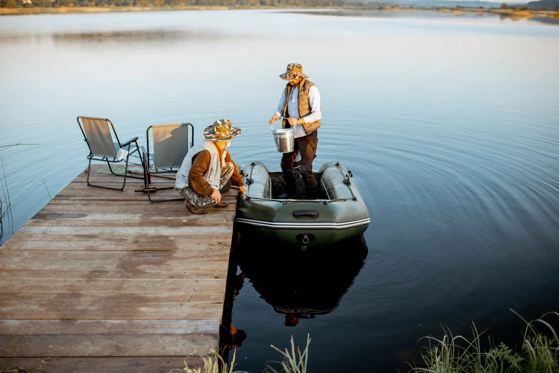 Two people at the end of a wooden dock on a still lake at dawn, one stepping into an inflatable boat.