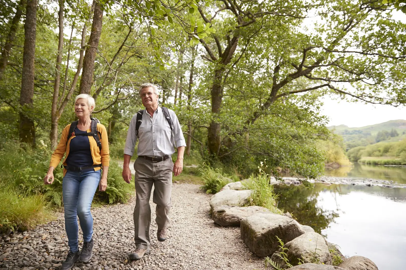 A couple walking a gravel riverside path under summer trees.
