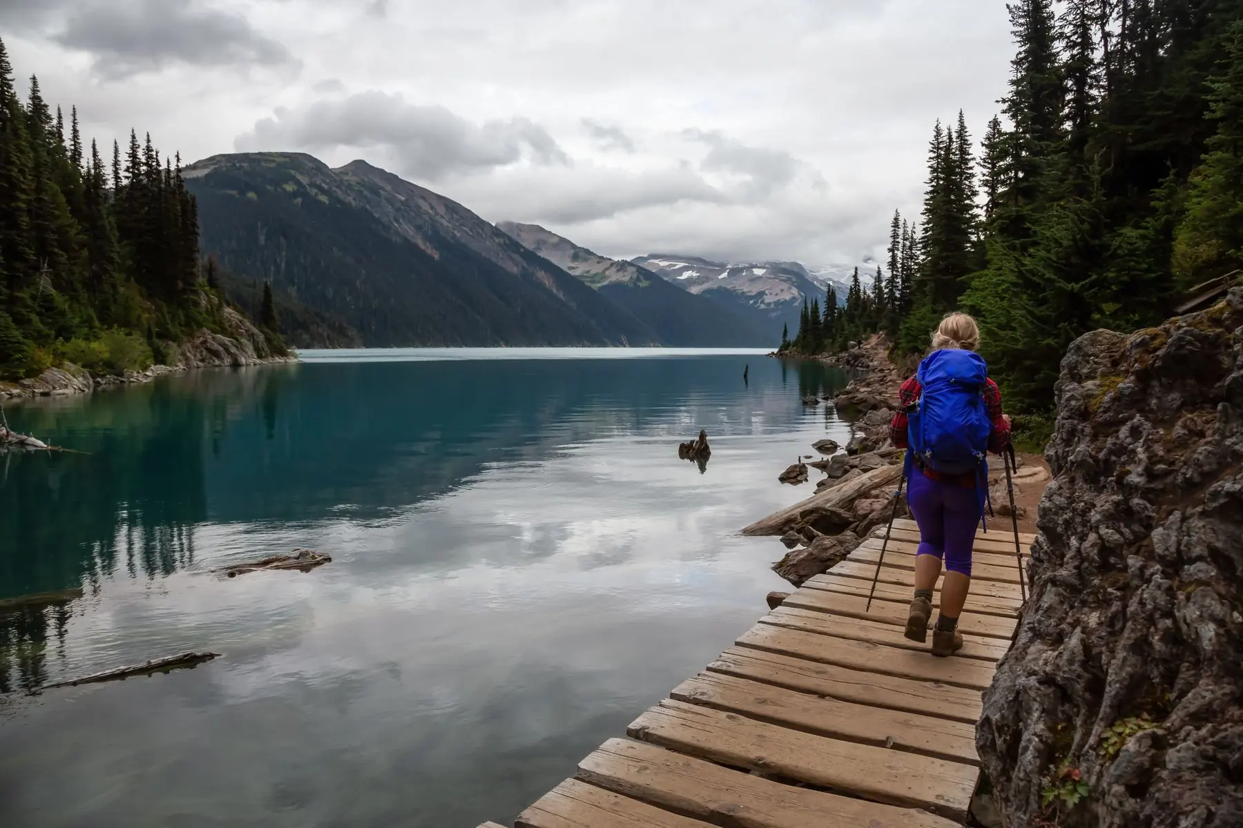 A hiker with a blue backpack walking a wooden boardwalk along a glacial lake.