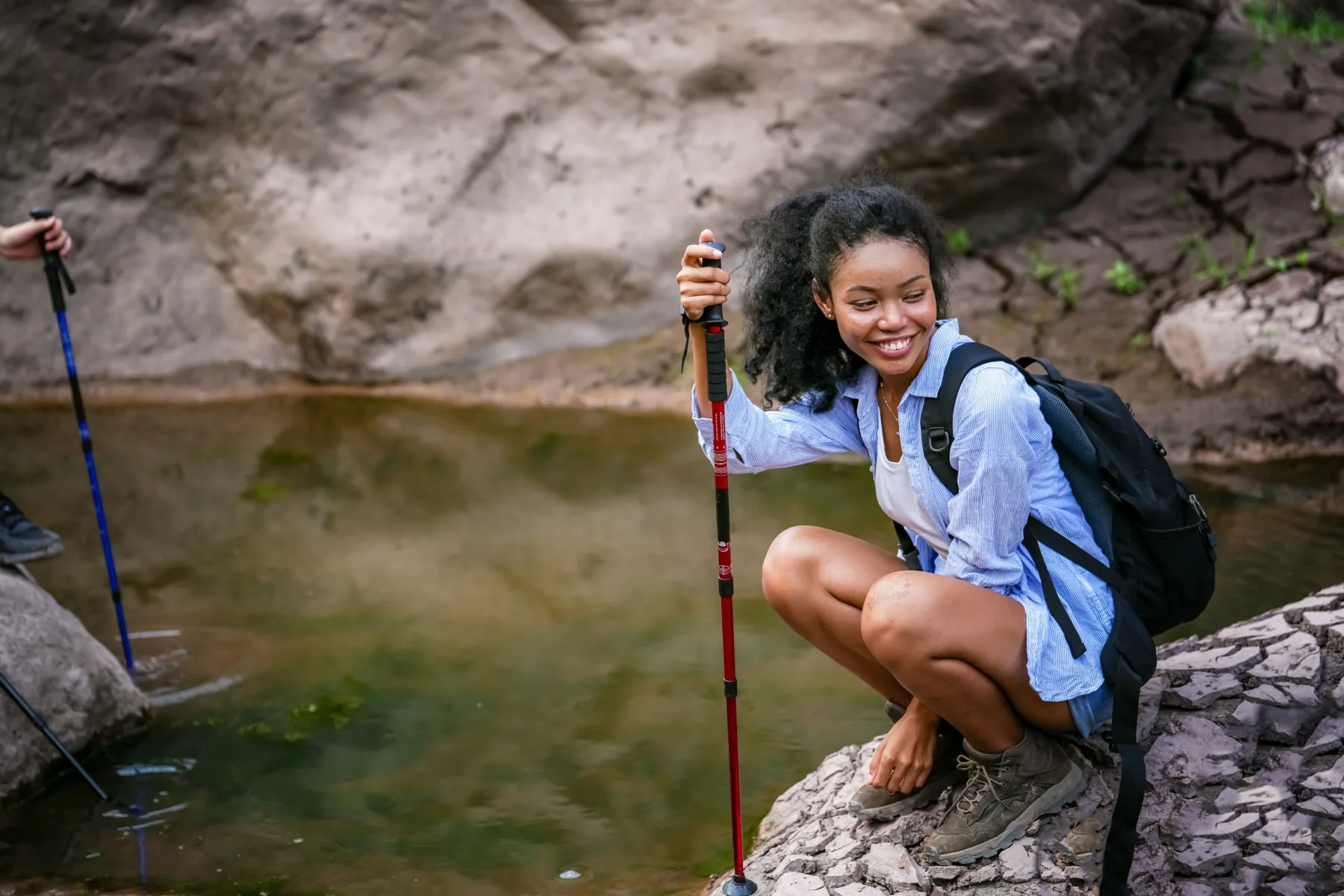 A hiker with trekking poles and a backpack crouching beside a clear pool along a rocky creek trail.