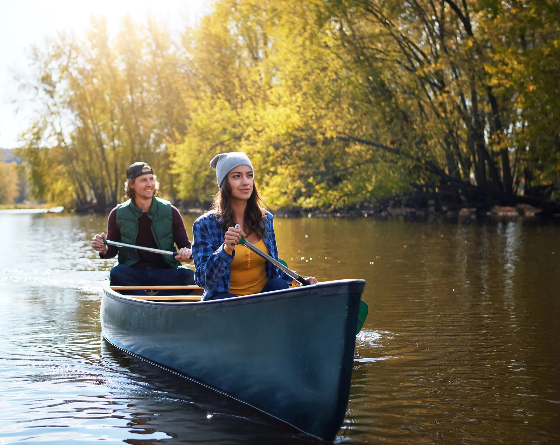 Two people paddling a green canoe down a calm river lined with autumn trees.
