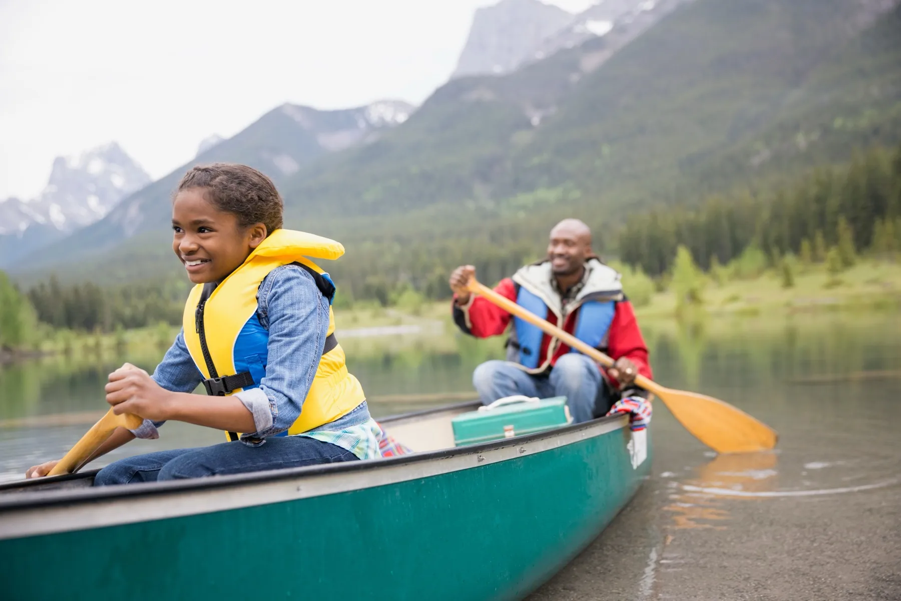 A girl in a life jacket paddling the bow of a green canoe on a mountain lake while her father paddles behind her.